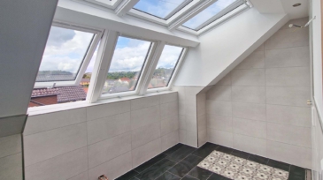 Ein Raum mit Fensterflächen auf dem Dach, große Fensterelemente in weißem Rahmen, mit Blick auf ein sonniges, bewölktes Land, beige Wandfliesen und schwarzem Boden mit einem area mit dekorativen Mustern, Kabel an der Wand.