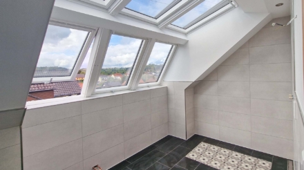 Ein Raum mit Fensterflächen auf dem Dach, große Fensterelemente in weißem Rahmen, mit Blick auf ein sonniges, bewölktes Land, beige Wandfliesen und schwarzem Boden mit einem area mit dekorativen Mustern, Kabel an der Wand.