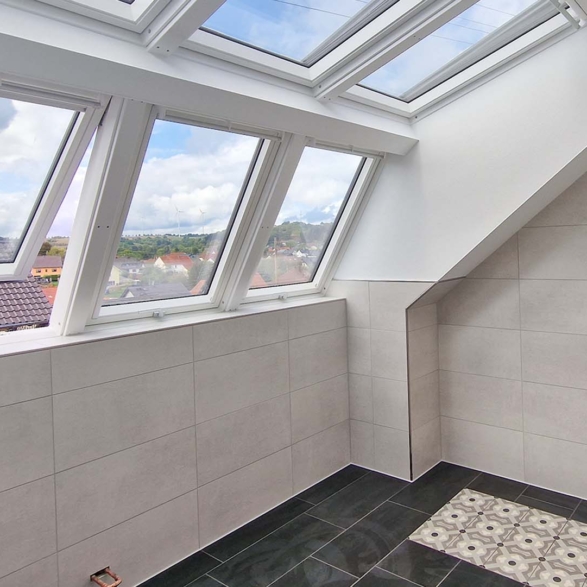 Ein Raum mit Fensterflächen auf dem Dach, große Fensterelemente in weißem Rahmen, mit Blick auf ein sonniges, bewölktes Land, beige Wandfliesen und schwarzem Boden mit einem area mit dekorativen Mustern, Kabel an der Wand.