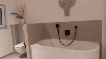 Ein Badezimmer mit einer Badewanne, an der eine schwarze Brausedusche und ein Wasserhahn angebracht sind, sowie eine weiße Toilette, ein Fenster mit einem kleinen Gegenstand auf dem Fensterbrett und eine Wanddekoration aus Makramé.