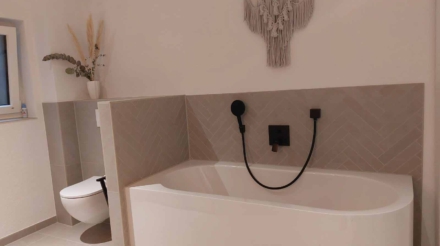 Ein Badezimmer mit einer Badewanne, an der eine schwarze Brausedusche und ein Wasserhahn angebracht sind, sowie eine weiße Toilette, ein Fenster mit einem kleinen Gegenstand auf dem Fensterbrett und eine Wanddekoration aus Makramé.