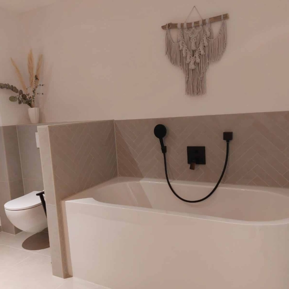 Ein Badezimmer mit einer Badewanne, an der eine schwarze Brausedusche und ein Wasserhahn angebracht sind, sowie eine weiße Toilette, ein Fenster mit einem kleinen Gegenstand auf dem Fensterbrett und eine Wanddekoration aus Makramé.