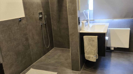 Ein modernes Badezimmer mit einer duschkabine, weißen toilette, einem weißen waschbecken mit schwarzem Unterschrank, einem Handtuch an der Tuchstange, einer Handseife im Seifenspender, einem großen Fenster mit digitalem Rolladen, grauen Fliesen an Wänden und Boden, und mehreren Deckenleuchten.