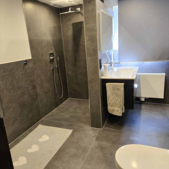 Ein modernes Badezimmer mit einer duschkabine, weißen toilette, einem weißen waschbecken mit schwarzem Unterschrank, einem Handtuch an der Tuchstange, einer Handseife im Seifenspender, einem großen Fenster mit digitalem Rolladen, grauen Fliesen an Wänden und Boden, und mehreren Deckenleuchten.