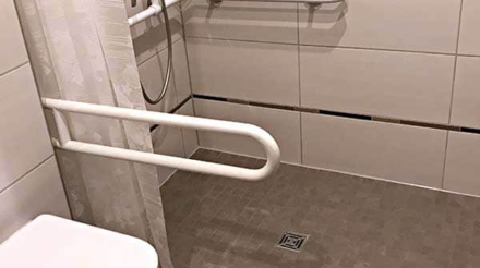 Ein barrierefreies Badezimmer mit einer steinernen Toilette, Handläufen an den Wänden, Duschutensilien auf einem Wandkorb und einer geteilten Dusche mit einem Wasserabfluss im Boden.