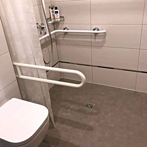 Ein barrierefreies Badezimmer mit einer steinernen Toilette, Handläufen an den Wänden, Duschutensilien auf einem Wandkorb und einer geteilten Dusche mit einem Wasserabfluss im Boden.