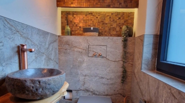 Innenansicht eines stilvoll gestalteten Badezimmers mit einer runden Waschschüssel aus rauem Naturstein, einem modernen grauen WC, einem Holzregal mit Handtuch und Deko, einer Wand aus Marmor und einer Holzfrontwand im Hintergrund, sowie einem großen Fenster auf der rechten Seite.