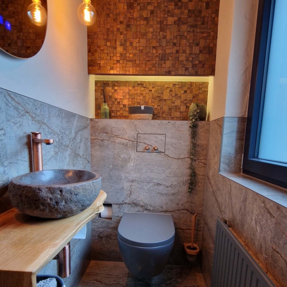 Innenansicht eines stilvoll gestalteten Badezimmers mit einer runden Waschschüssel aus rauem Naturstein, einem modernen grauen WC, einem Holzregal mit Handtuch und Deko, einer Wand aus Marmor und einer Holzfrontwand im Hintergrund, sowie einem großen Fenster auf der rechten Seite.