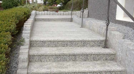 Eine Steinrampe mit mehreren Stufen, umgeben von grünen Sträuchern und einer Schrankenmauer aus Steinen. An der Rampe sind handgefertigte Metallgeländer angebracht sowie schmaler Kies zu sehen.