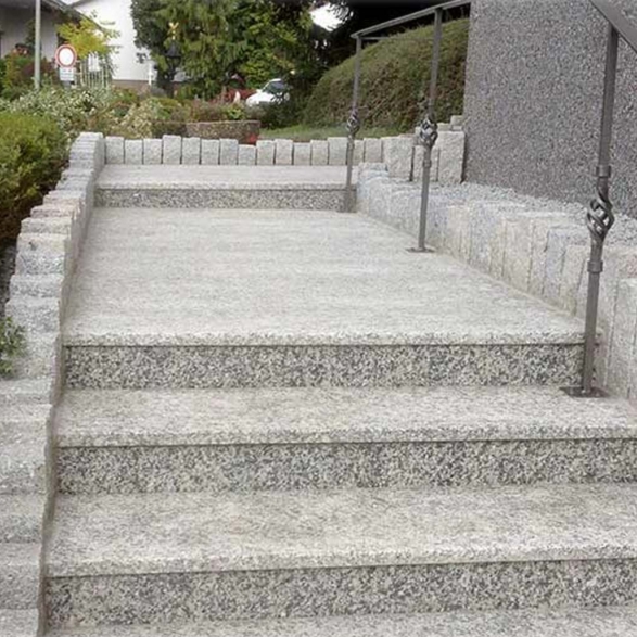 Eine Steinrampe mit mehreren Stufen, umgeben von grünen Sträuchern und einer Schrankenmauer aus Steinen. An der Rampe sind handgefertigte Metallgeländer angebracht sowie schmaler Kies zu sehen.