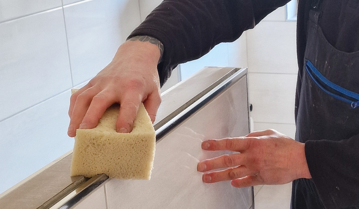 Eine Person reinigt eine Fliesenwand mit einem Schwamm, trägt eine schwarze Arbeitsjacke und hat einen Tattoo auf dem Handgelenk.
