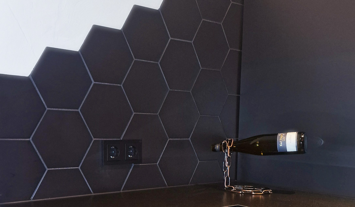 Eine dunkle Flasche mit einem Kettenverschluss steht gegen eine dunkle Wand mit Hexagon-Fliesen, neben einer doppelt steckdose, im Hintergrund eine weiße Wand.