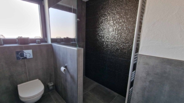 Ein Badezimmer mit einer Toilette, einem grauen Fliesenwand, einem Fenster mit drei kleinen Dekogläsern auf der Fensterbank und einer schwarzen Wand mit strukturierter Oberfläche im Duschbereich.