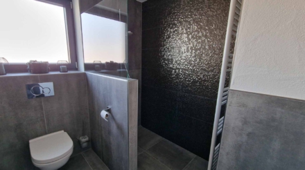 Ein Badezimmer mit einer Toilette, einem grauen Fliesenwand, einem Fenster mit drei kleinen Dekogläsern auf der Fensterbank und einer schwarzen Wand mit strukturierter Oberfläche im Duschbereich.