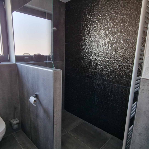 Ein Badezimmer mit einer Toilette, einem grauen Fliesenwand, einem Fenster mit drei kleinen Dekogläsern auf der Fensterbank und einer schwarzen Wand mit strukturierter Oberfläche im Duschbereich.