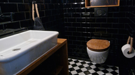 Ein Badezimmer mit schwarzen Wandfliesen, einem weißen Waschbecken auf einem Holzunterschrank, einem wandhängenden WC mit Holzdeckel, schwarzem Fliesenboden mit weißem Muster, einem runden Spiegel und einem Wandbild mit einem weiblichen Kopf, alles im modernen Design.