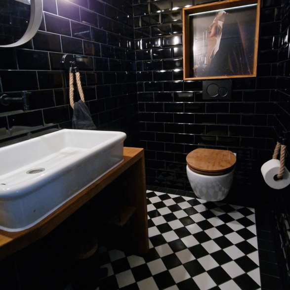 Ein Badezimmer mit schwarzen Wandfliesen, einem weißen Waschbecken auf einem Holzunterschrank, einem wandhängenden WC mit Holzdeckel, schwarzem Fliesenboden mit weißem Muster, einem runden Spiegel und einem Wandbild mit einem weiblichen Kopf, alles im modernen Design.