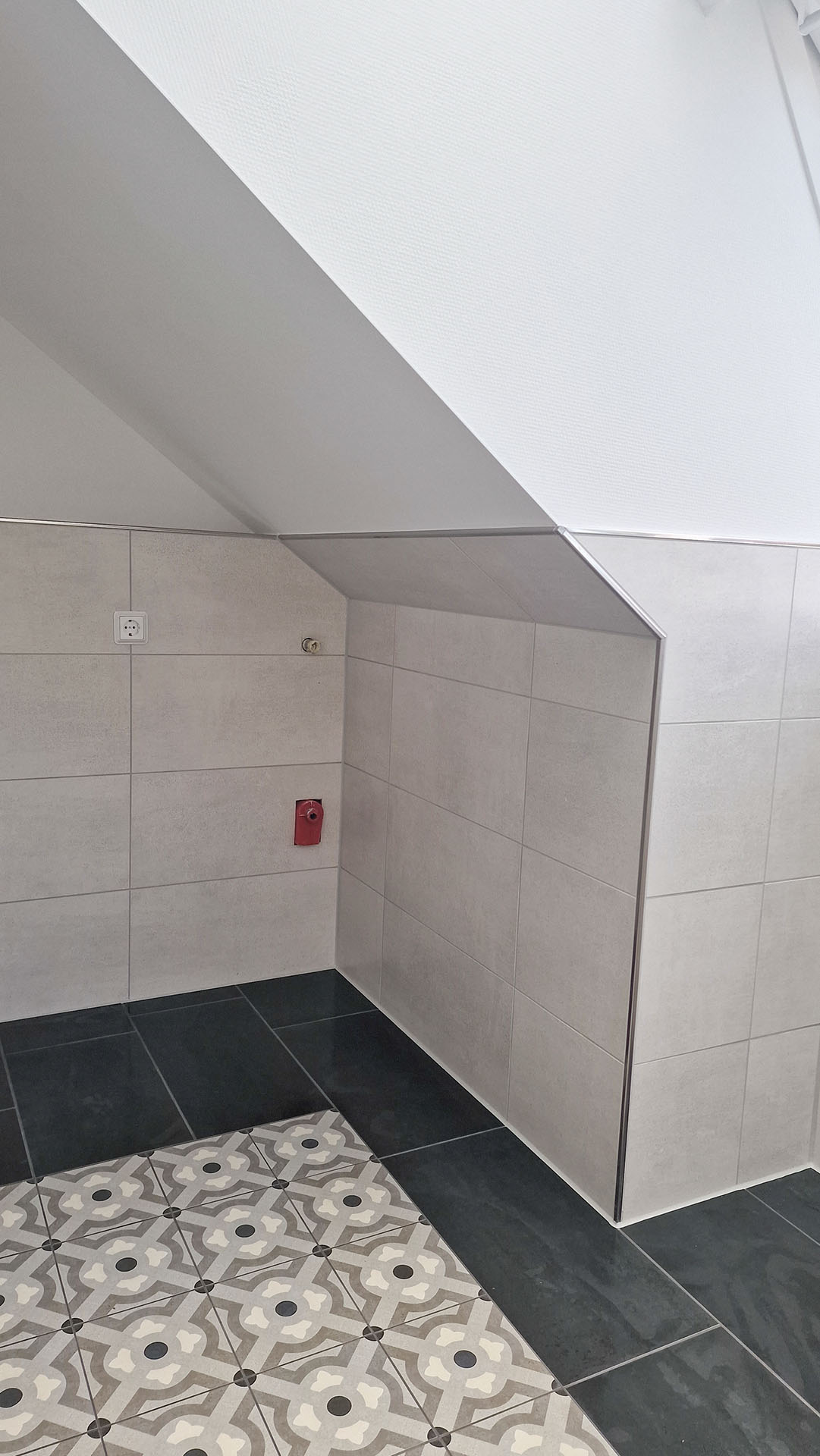Innenansicht eines Raums mit gemusterten Bodenfliesen und hellen Wandfliesen, einer roten Wandsteckdose und unvollständigen Installationen an der Wand unter einer abgehängten Decke.