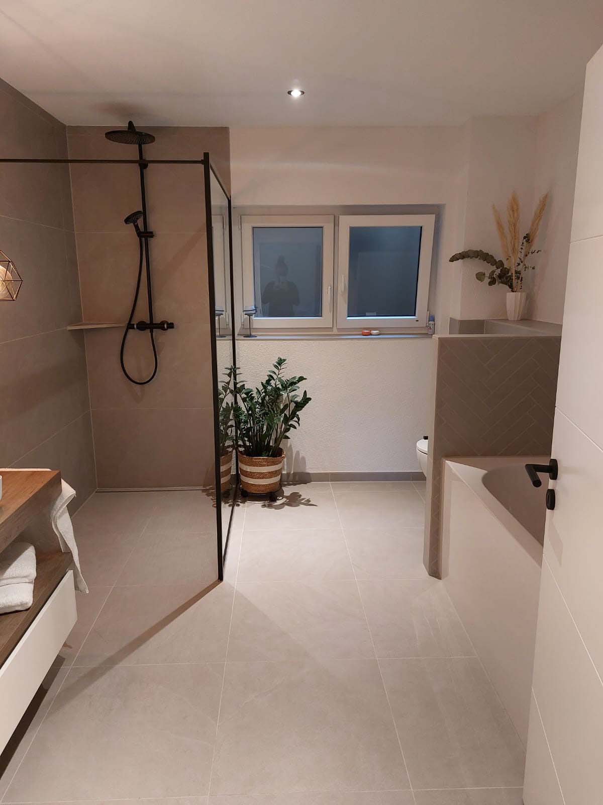 Ein modernes Badezimmer mit einer begehbaren Dusche, einer Pflanze in einem geflochtenen Korb, einem Waschbecken mit schwarzer Armatur und einem Fenster, das Tageslicht hereinlässt