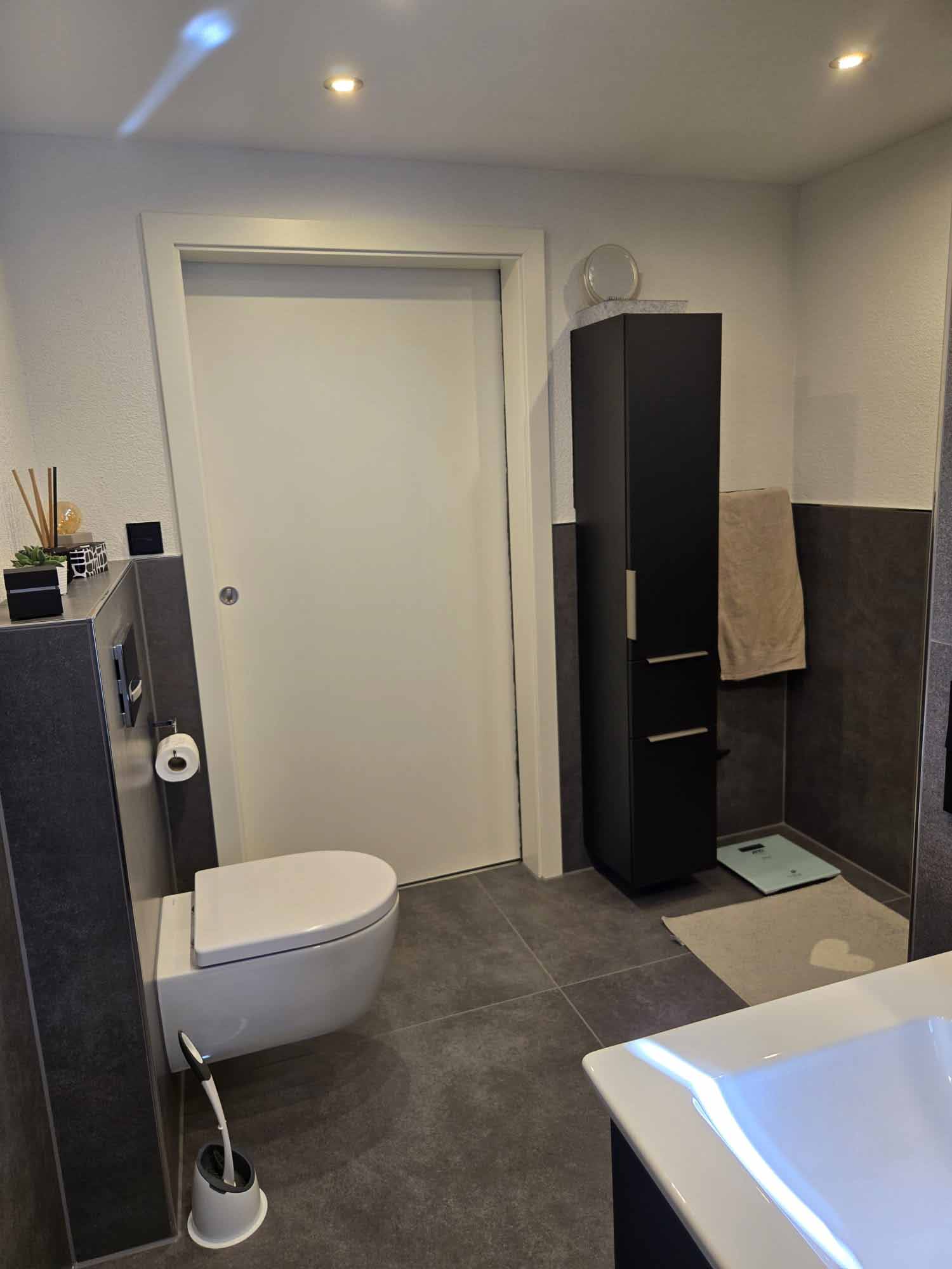 Ein Badezimmer mit Toilette, schwarzen Schränken, Handtüchern und einer digitalen Waage auf dem Boden, grau-weißen Wänden und Deckenleuchten.