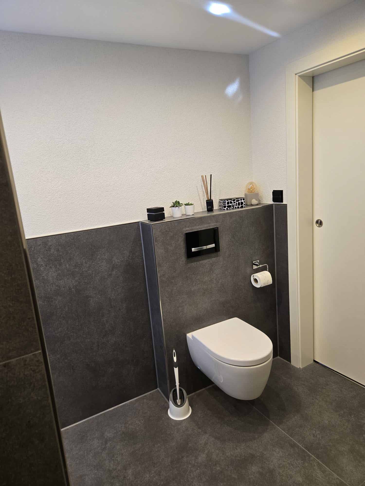 Ein modern ausgestattetes Badezimmer mit einer Wand-WC, grauen Fliesen und weißen Wänden, Dekorationsgegenständen auf einer Ablage, einem Toilettenpapierhalter und einem Toilettenbürstenständer.