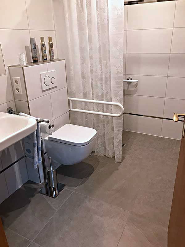 Ein modernes, helles Badezimmer mit einer Dusche, einem Waschbecken und einer Toilette. Die Wände sind mit weißen Fliesen verkleidet, der Boden ist grau gefliest. Neben der Toilette befindet sich ein Haltegriff, und auf dem Waschbecken stehen Pflegeprodukte.
