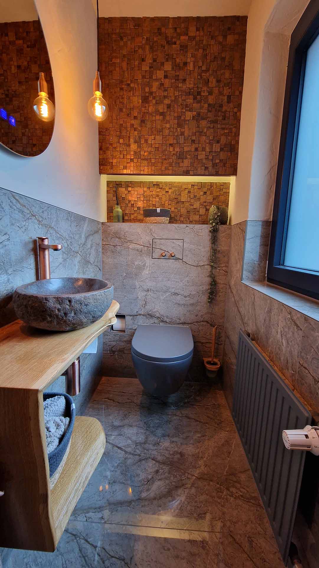 Ein modernes Badezimmer mit einer ovalen Fenster, einem Wand-WC, einer Steinwaschschüssel auf einem Holzregal, einer Wand aus Marmorfliesen, einer welligen weißen Wand, einer warm beleuchteten Holzlampe und einer braunen Ziegelwand im Hintergrund.