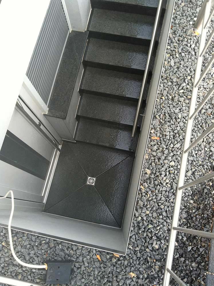Blick von oben auf eine schwarze Außentreppe aus Metall mit einem kleinen Podest und einem Entwässerungsrost in der Mitte, umgeben von grauem Kies, rechts ein Metallgeländer und unten ein weißes Kabel mit schwarzem Stecker.