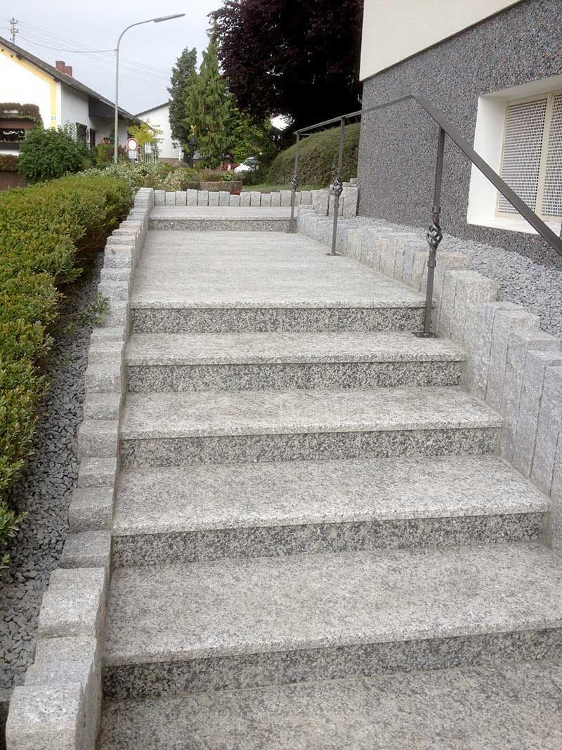 Die Treppe aus grauem Granit hat mehrere Stufen und ist mit einem metallenen Handlauf versehen. An den Seiten sind niedrige, graue Steinmauern. Im Hintergrund sind Bäume und einige Gebäude sichtbar.