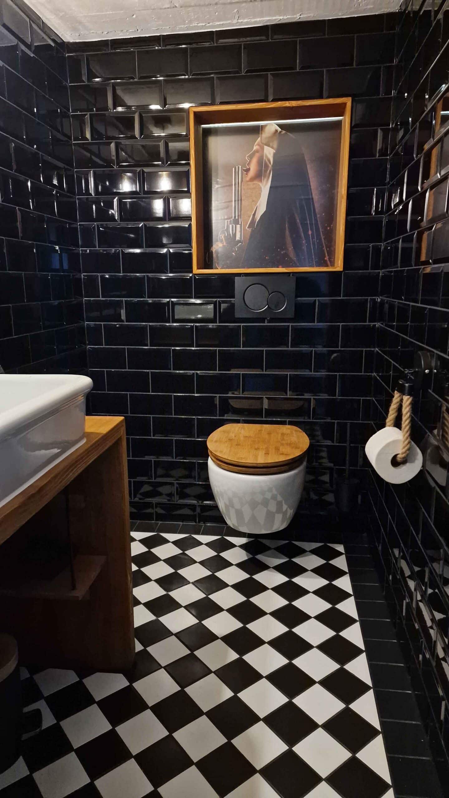 Ein Badezimmer mit schwarzen Wandfliesen, schwarz-weiße Fliesen auf dem Boden, einer weißen Toilette mit Holzdeckel, einem weißen Waschtisch aus Keramik und einem Bilderrahmen mit einem Foto, das eine Frau mit einem Revolver zeigt, hängt an der Wand.